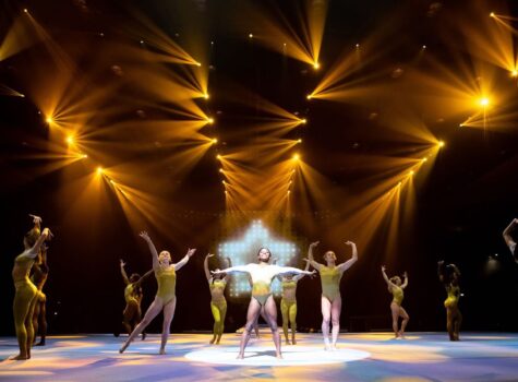 a group of gynamstic on stage with gold outfits, and lights in the shape of a star behind the center