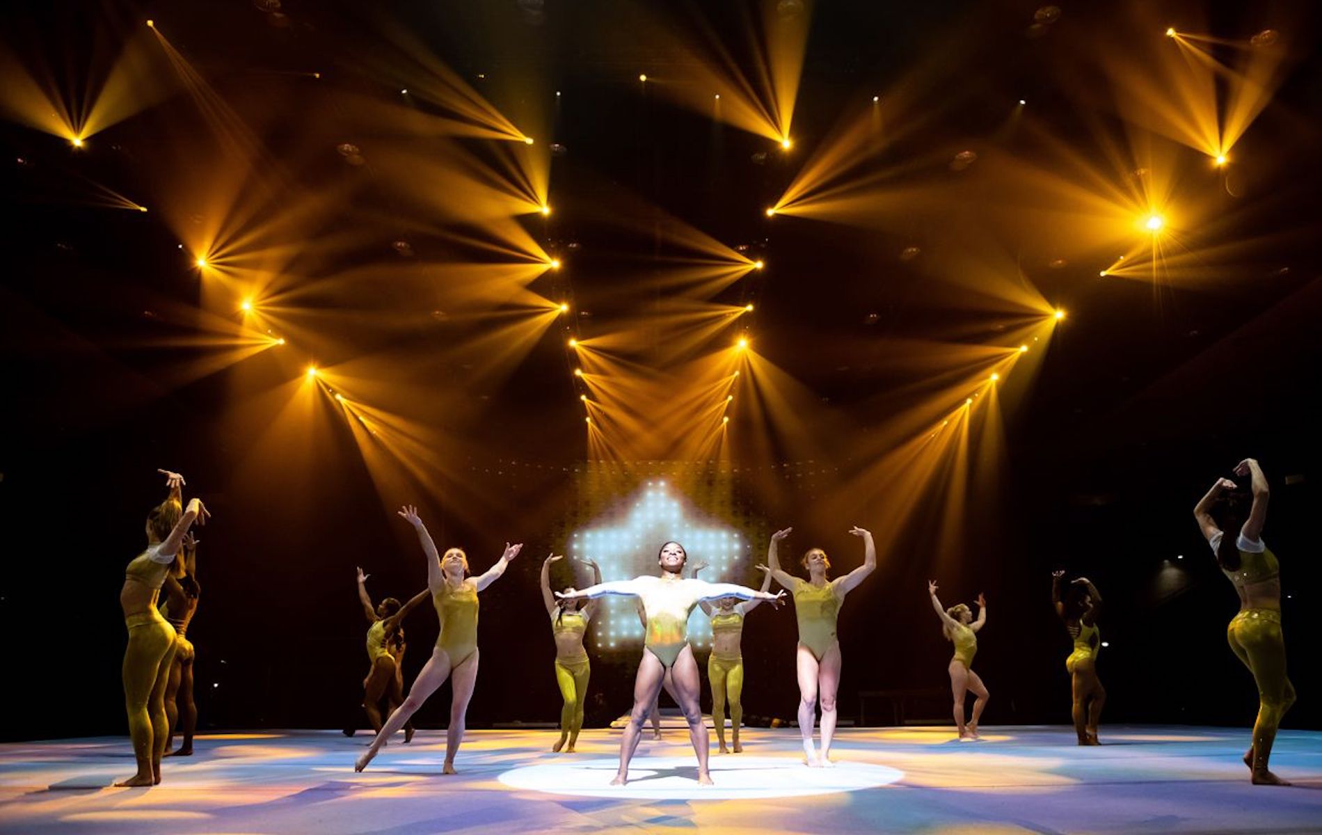 a group of gynamstic on stage with gold outfits, and lights in the shape of a star behind the center