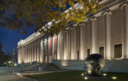 Museum with white columns at night with tree and gaint black state of a head