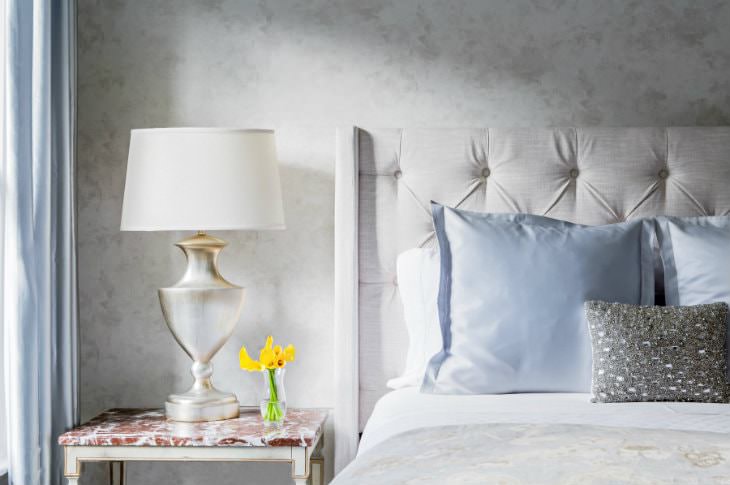 Bed with beige tufted headboard, white sheets and blue pillows, white nightstand with white-washed gold lamp and yellow flowers