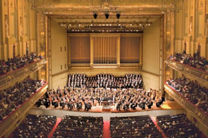 Elegant and elaborate auditorium with gold walls and tall ceilings filled with an orchestra and audience