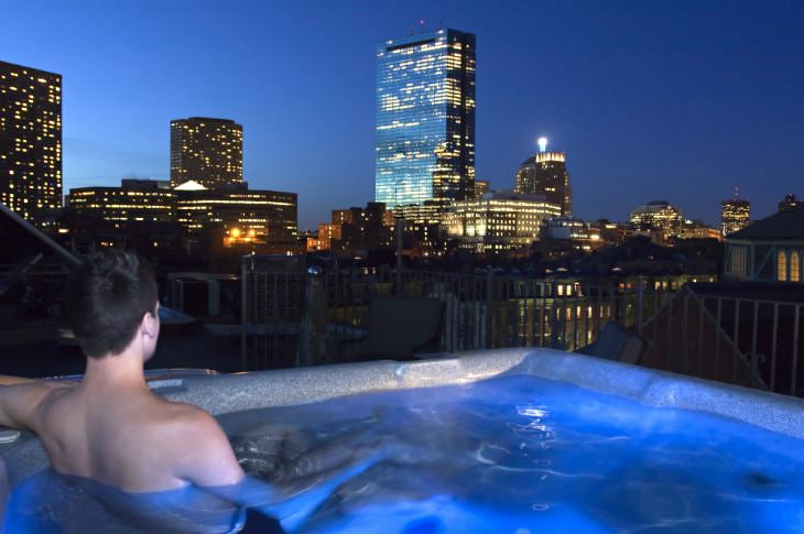 Man in roof top hot tub overlooking the dark blue sky and twinkling city at night
