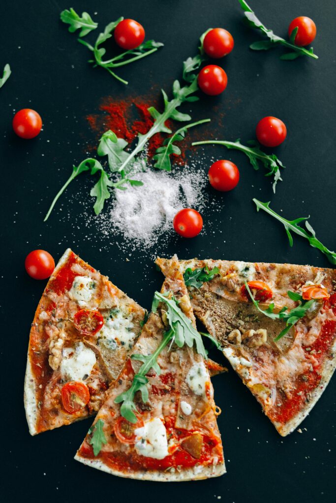 Three pieces of pizza on a black board with red tomatoes and green leafy  arugula scattered and a small pile of grated white cheese in center